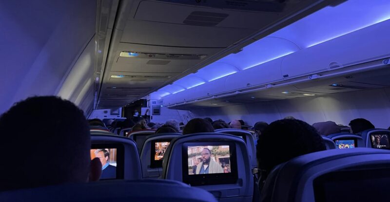 Delta 757-200 cabin interior with blue LED lighting. Passengers are throughout the aircraft. The view is from the back of the aircraft showing the IFE screens at each seat.