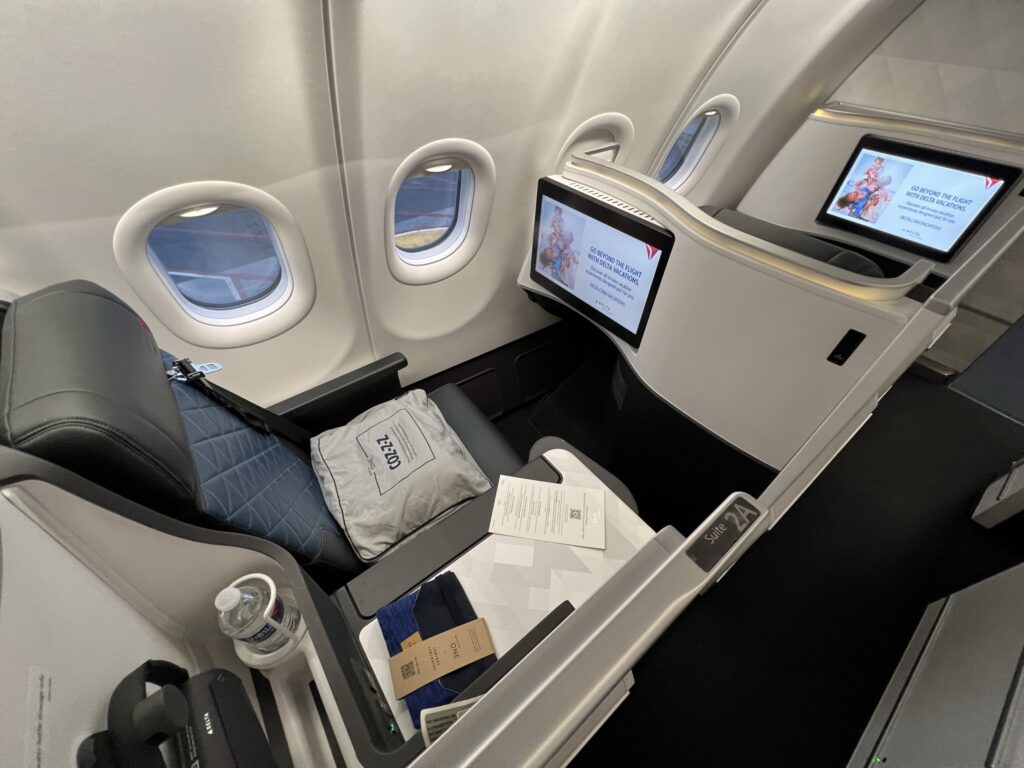 Delta A330-900neo suite is pictured here. The seat is blue with a much darker blue headrest and the outer thermoplastics and details are light grey, Headphones are hanging in the suite and bedding is waiting for the passenger on the seat. 