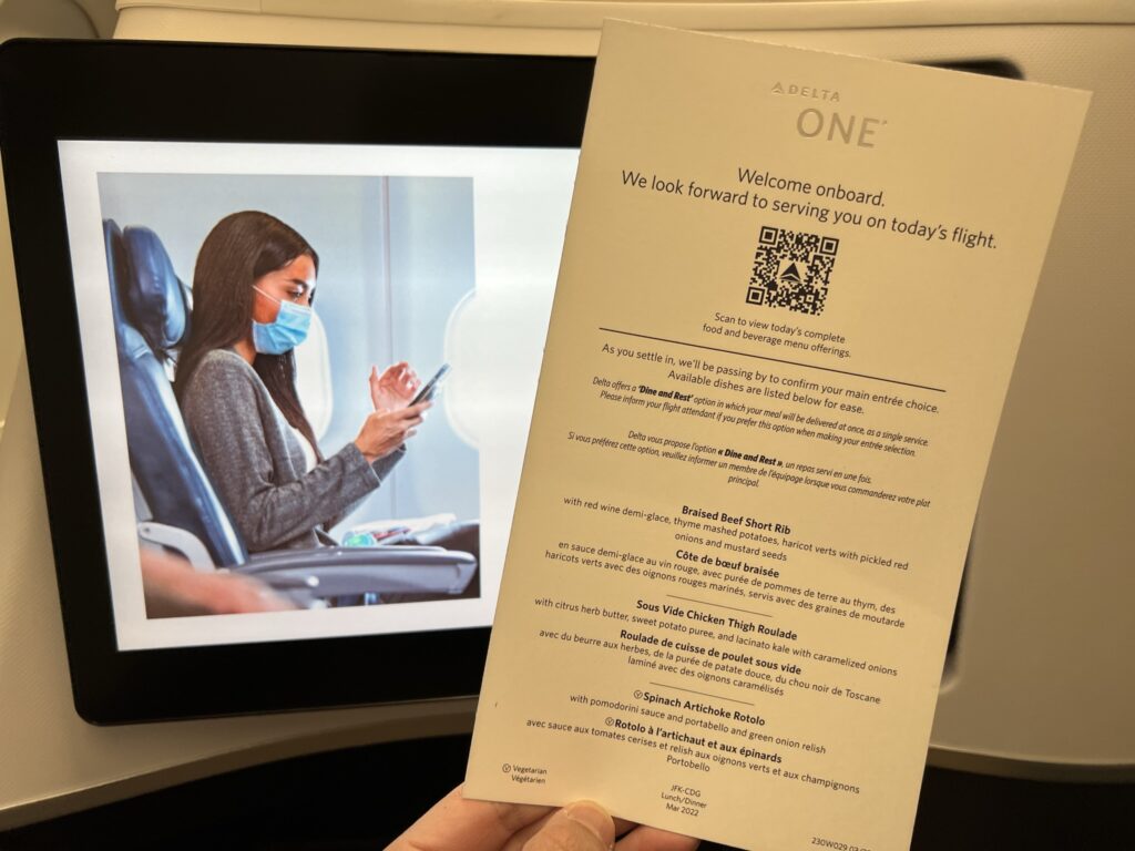 A man is holding up Delta's menu in front of the IFE screen to show the QR code that allows access to more options. 