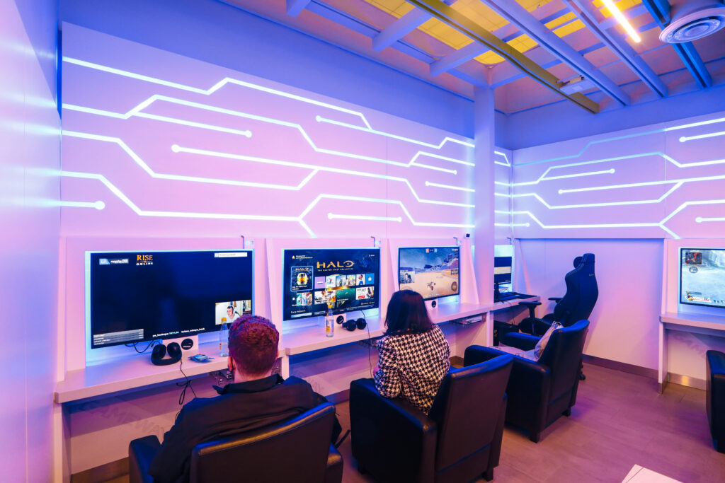 Passengers that are waiting for flights to depart sit playing games in Gameway's premium gaming lounge at LAX. 3 systems are set up in a row with comfortable chairs. Above the television screens the walls are decorated to look like a circuit board.