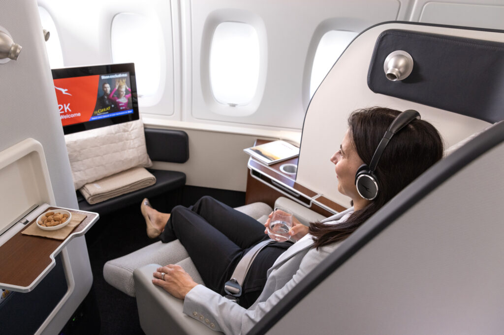 A female passenger is sitting in the Qantas first class seat with a drink in hand. She is wearing headphones as she watches the IFE screen.