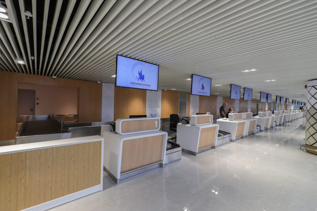 Air Marakanda checkin counters are pictured here. There each have large digital screens to display information. and the counters are made up of light pine wood panels and white accents.