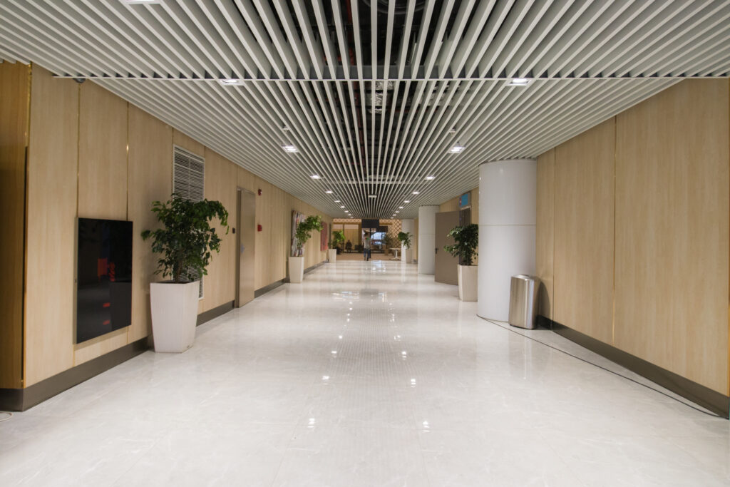 A long corridor in the Samarkand International Airport. The ceiling is wood paneling and the walls are all made up of light pine wood panels. There is a bright white floor and large plants in large white planters.