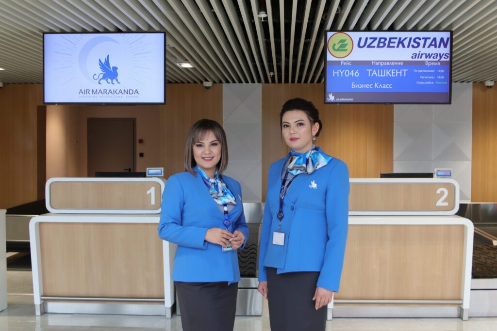 Two female Air Marakanda crew members are standing in front of the checkin counters. They are wearing baby blue jackets .