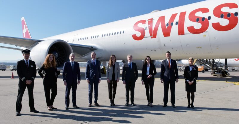 A group of executives are standing in a row in front of a Swiss aircraft on a sunny day.