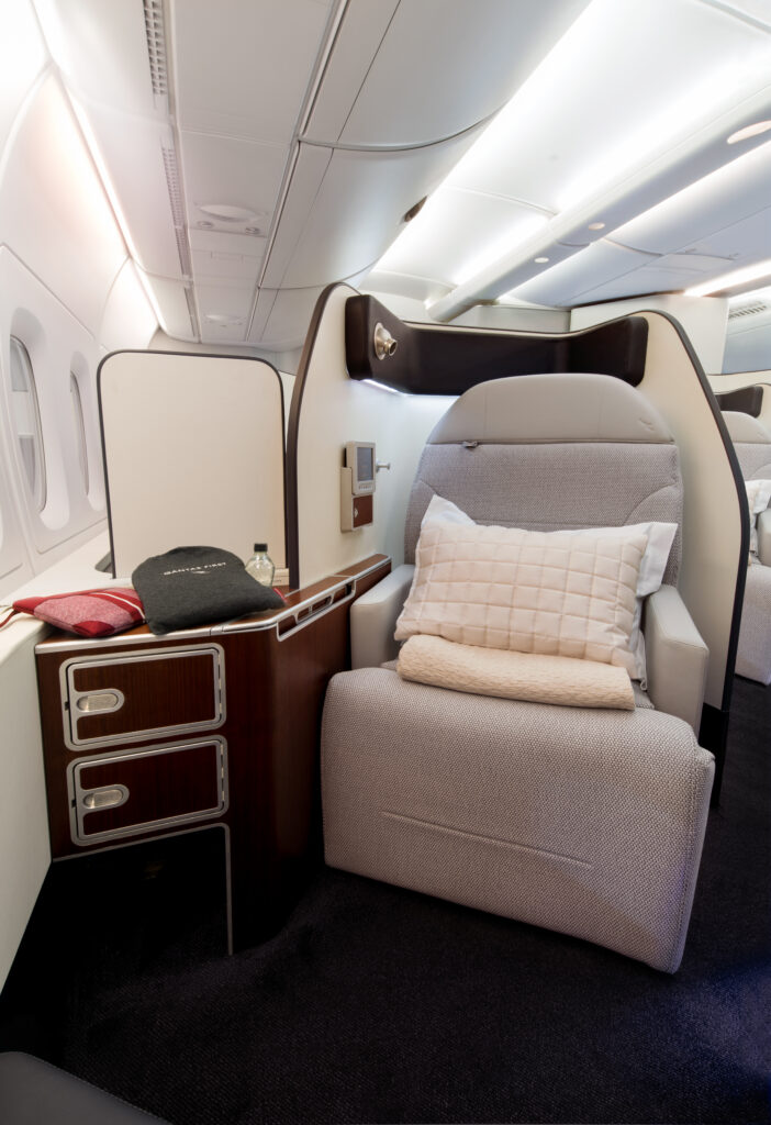 Qantas first class seat pictured here head on. The seat is light grey with a large white pillow set on top. The thermoplastic shell is white and storage areas are chocolate brown with wood finish.