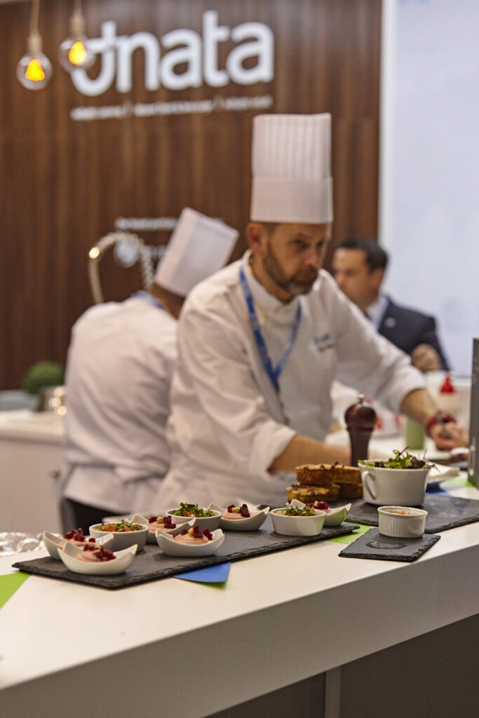 A chef at the dnata stand is standing in front of a long table full of samples of food.