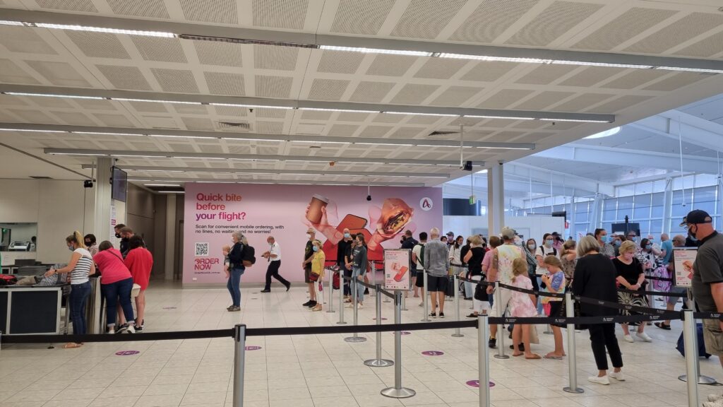 A large Servy advertisement is seen on the wall of an airport. A line up of people are waiting at check in.