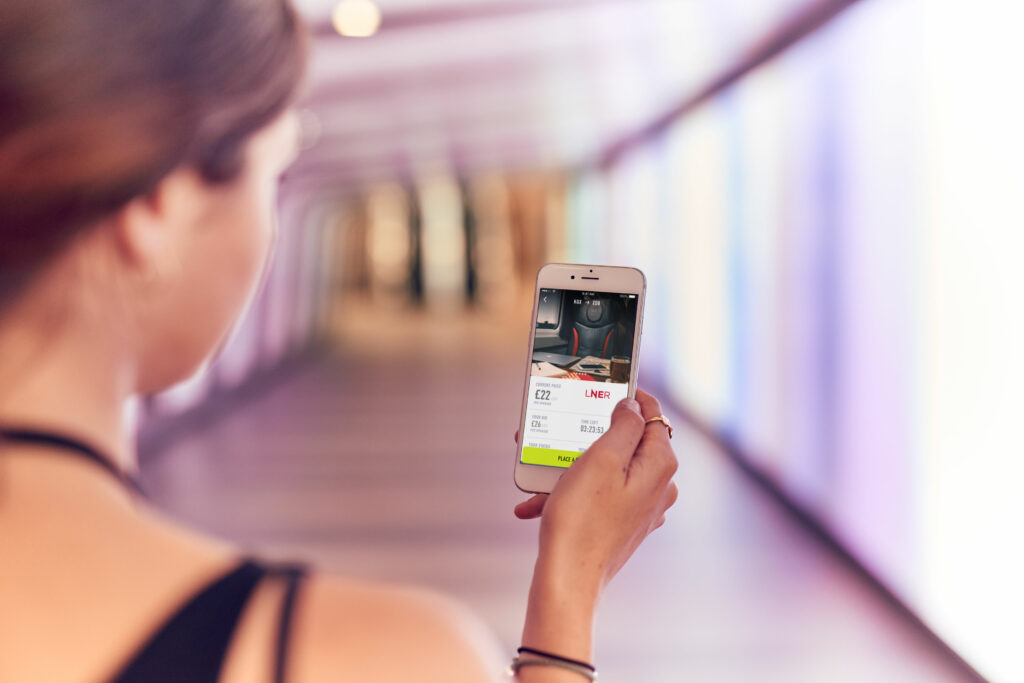 A woman is holding her mobile device in the train station. The device is currently displaying her bid for a first class seat upgrade.