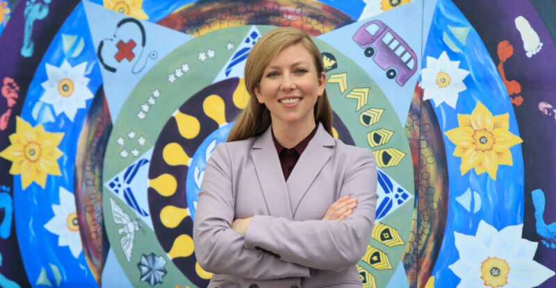 Tiffany Black stands in front of the mural. The mural is a colorful piece, with circles building up to add more detail to the art as it gets larger and larger.