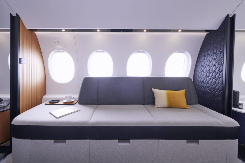 Dassault Falcon 10X sofa which can be converted into a bed. The bed runs the length of 4 aircraft windows and is light and dark grey with a yellow pillow.