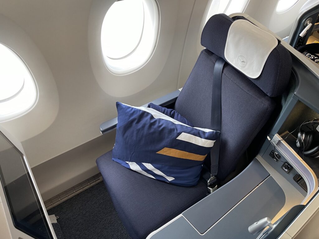 Lufthansa A350 Vantage XL business class seat. The blue seat has a white headrest cover and a blue pillow with orange and white stripes.