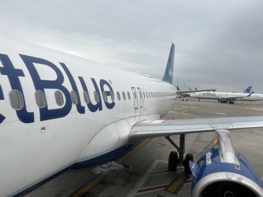 The side of a JetBlue A320 aircraft. The aircraft is white with a blue engine and tail. 