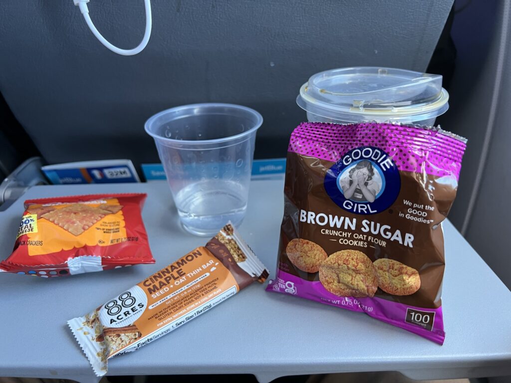 A bag of cookies, crackers and a cinnamon bar are displayed on the aircraft tray table.