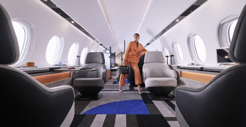 Dassault Falcon 10X interior. A woman in a suit leans on one of 4 visible chairs. A geometric carpet in grey, black and blue gives the space a modern feel.