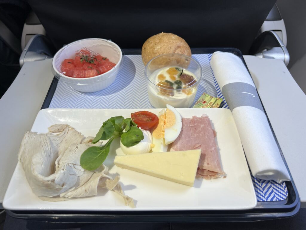 A piece of cheese, some processed meat and a egg sit on a plate on the tray table aboard this British Airways A320neo flight