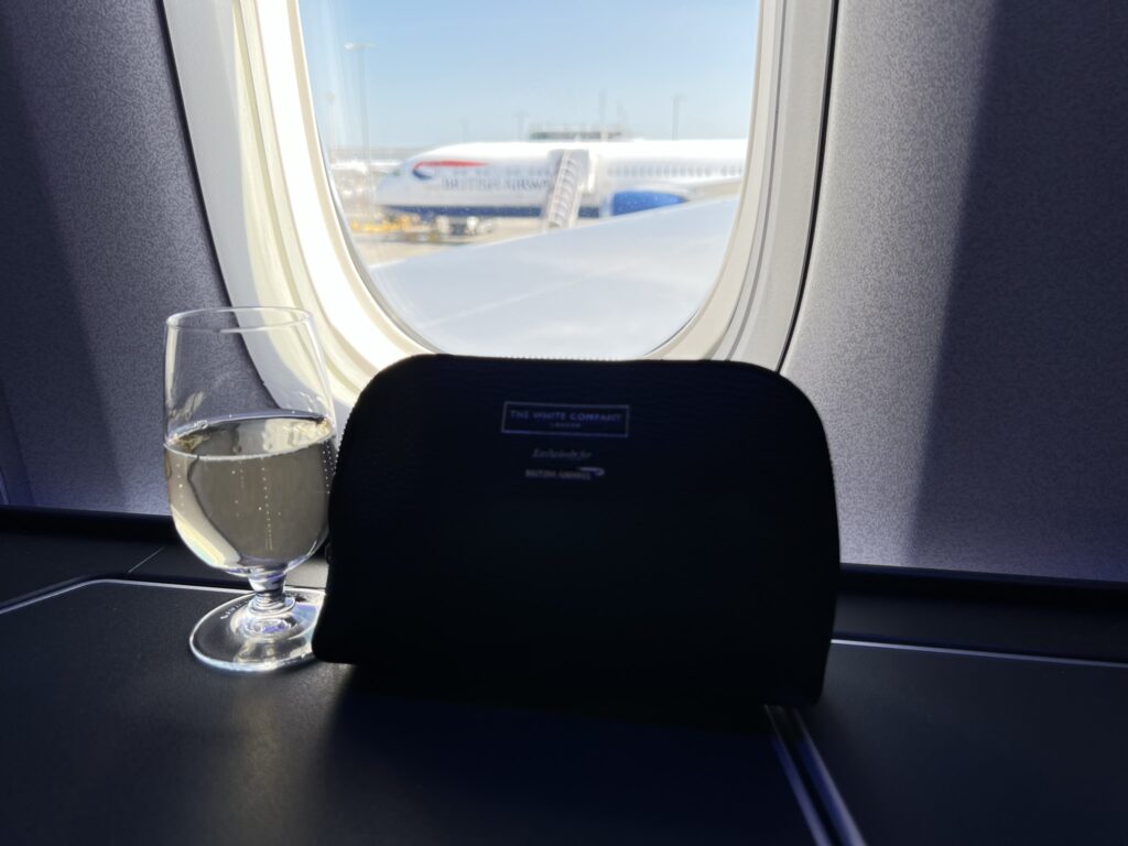 A glass of champers and an amenity kit sit side-by-side. A BA A380 is seen outside of the window.