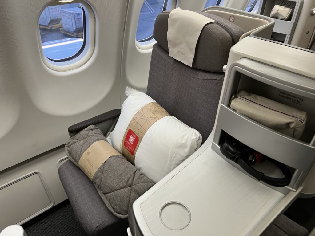 Close-up photo of a true window seat in business aboard the Iberia A330-200. The seat is all grey with a white pillow and grey blanket sitting on top.