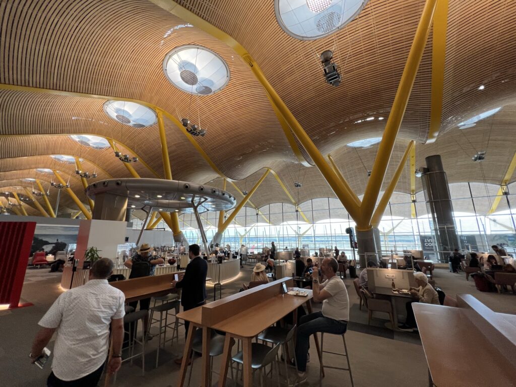 Iberia's spacious airport lounge at Madrid, with high ceilings and interesting light fixtures. Various seating options are shown, as passengers buzz about