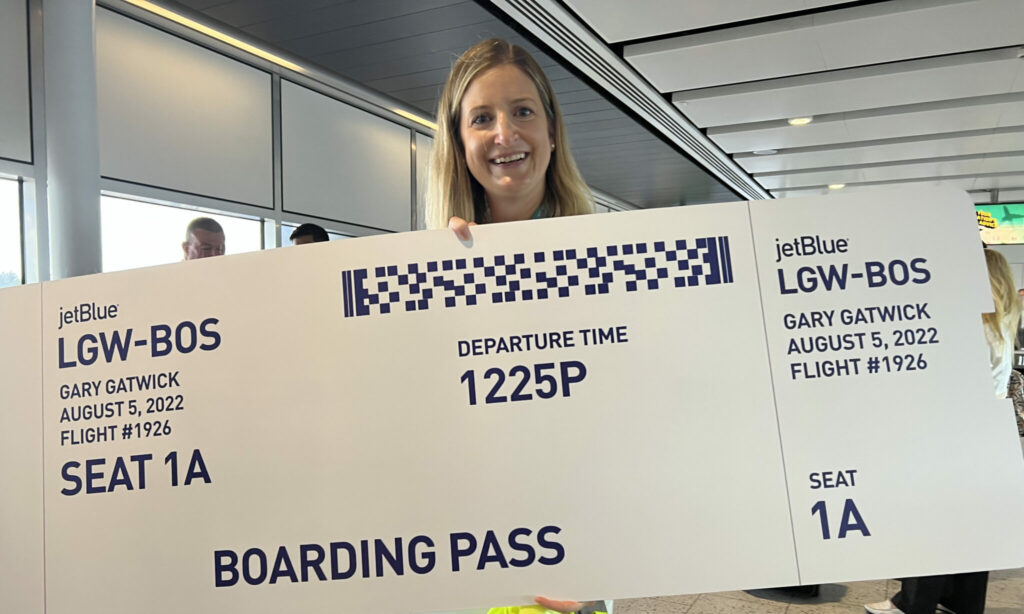 A woman is holding up a giant JetBlue boarding pass to celebrate the LGW-BOS inaugural