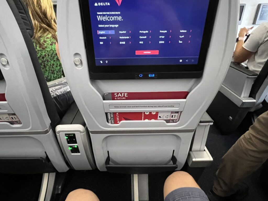 The author is seated in a Delta Premium Select seat. His legs are in view, showing ample legroom. An in-seat IFE screen is also in this shot.