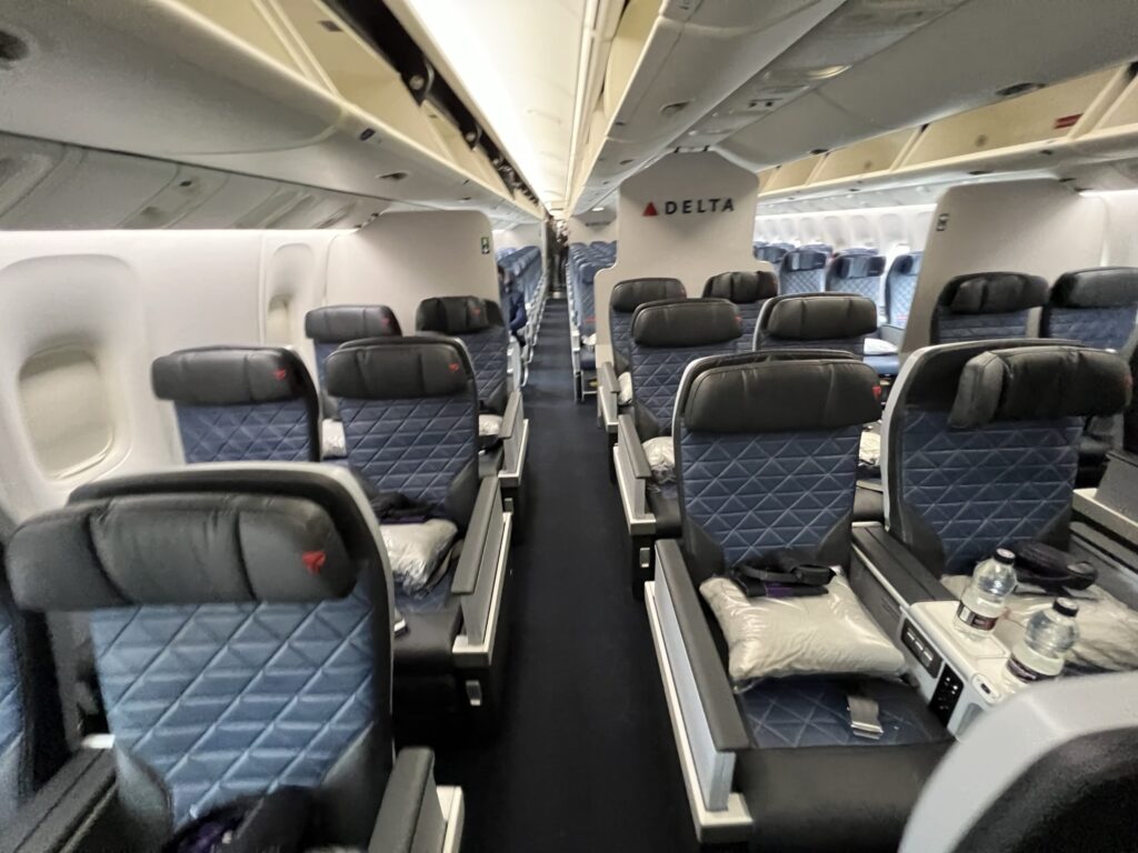 A view of the Delta Premium Select cabin. The seats are in a 2-2-2 configuration. They are dark blue with the red delta check on the headrest. Water bottles and pillows are displayed with the seats.