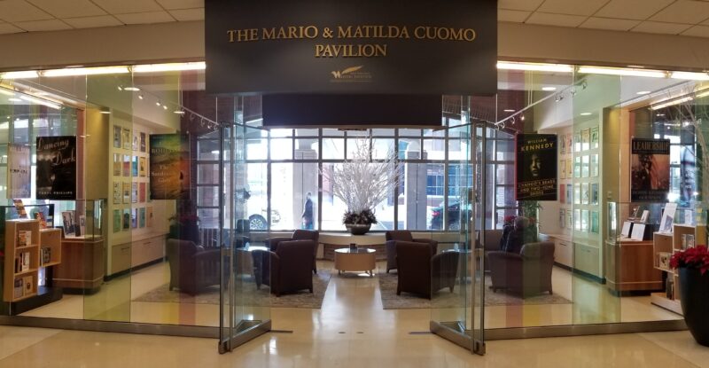 A large glass entry way into the Albany International Airport reading lounge. A sign above the entry way says "The Mario and Matilda Cuomo Pavilion"