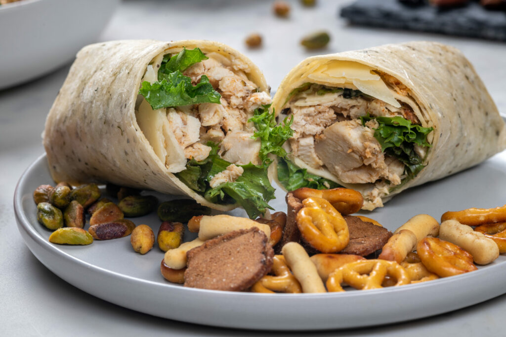 A chicken wrap is displayed on a white plate with pretzels and other snack bites