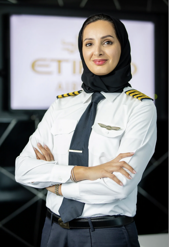 Captain Aisha Al Mansoori is posing for a photo with the Etihad logo behind her