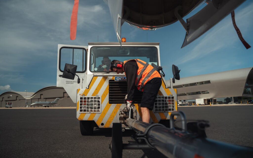 A man hooks up an aircraft to a tug machine on the ground at Farnborough Airport.