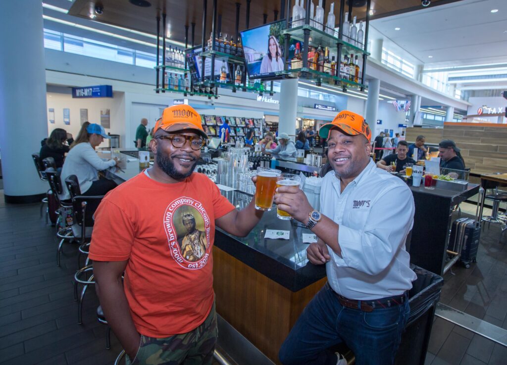 Two men are holding up beers at the airport bar.