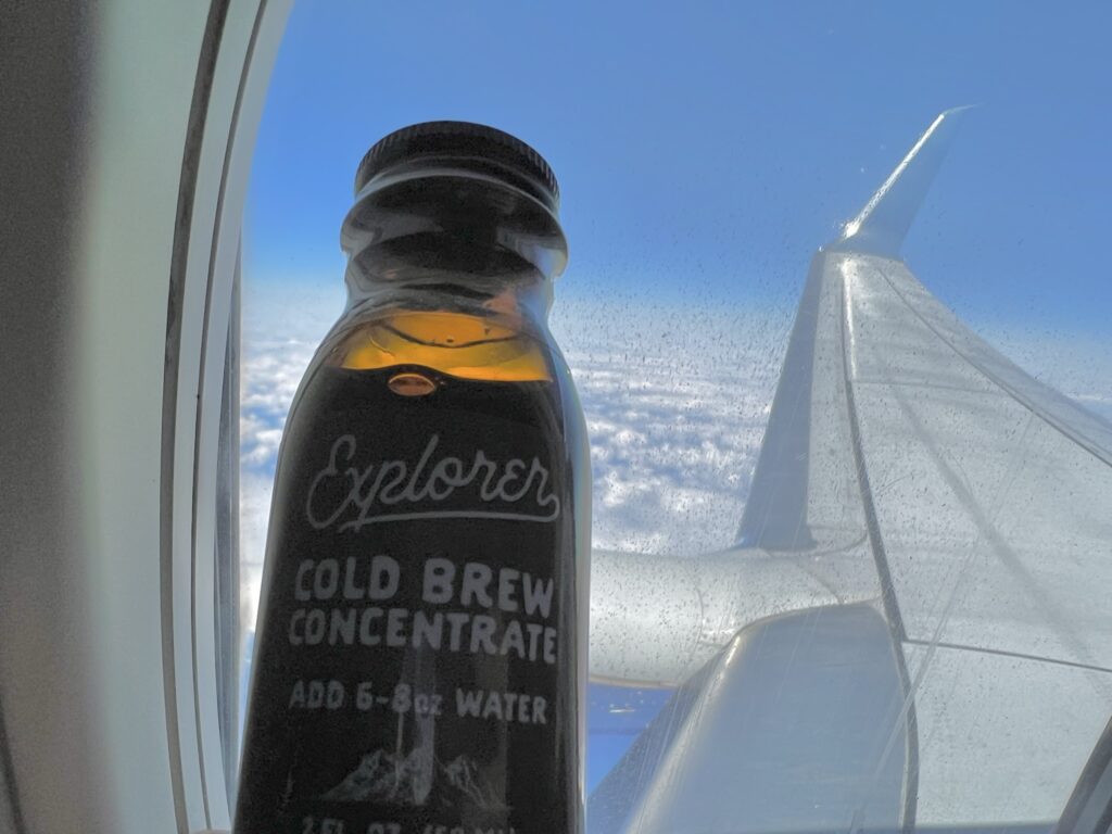 A passenger is holding up a cold brew bottle in front of the aircraft window.