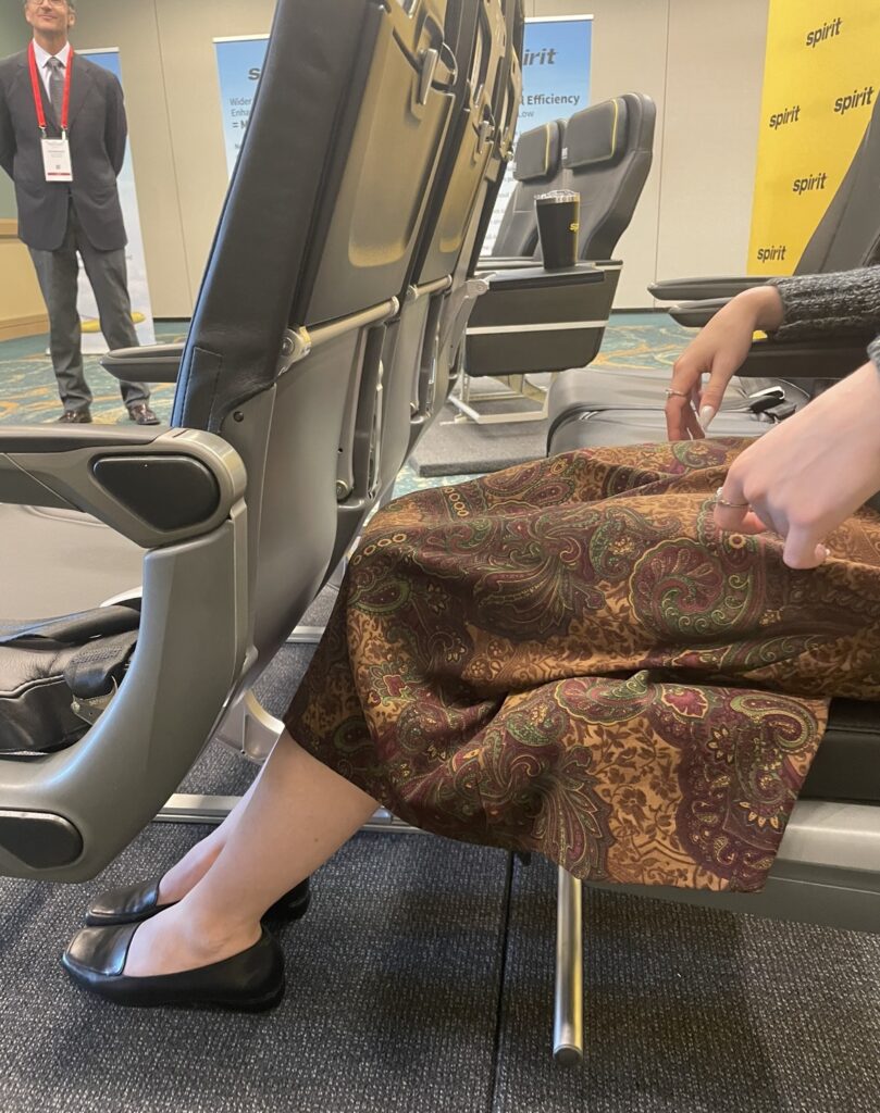 A young woman is sitting in the new Spirit Airlines economy class seat at the apex show demonstrating the leg room