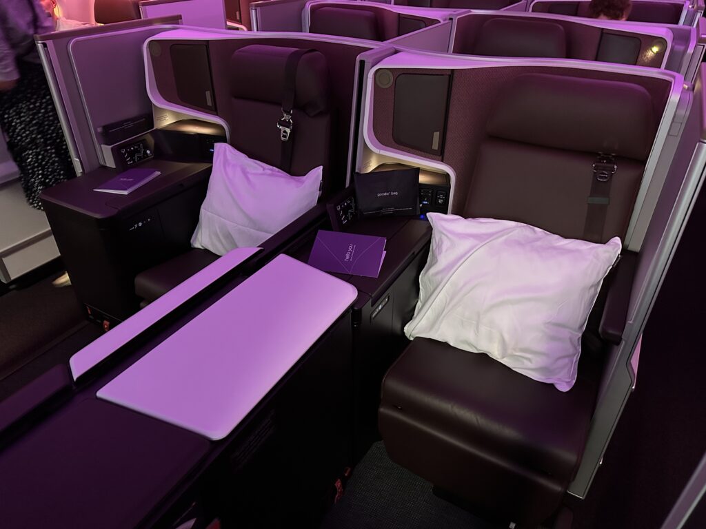 Virgin Retreat Suites are pictured. Two large seats, in dark brown covers, sit side-by-side. Purple LED lights cast a pretty glow on the surrounding furniture. A large white pillow sits on each seat.