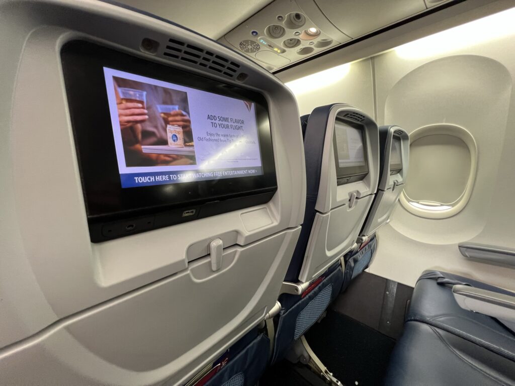 IFE screen is prominently in view, featuring an advertisement for an onboard beverage 