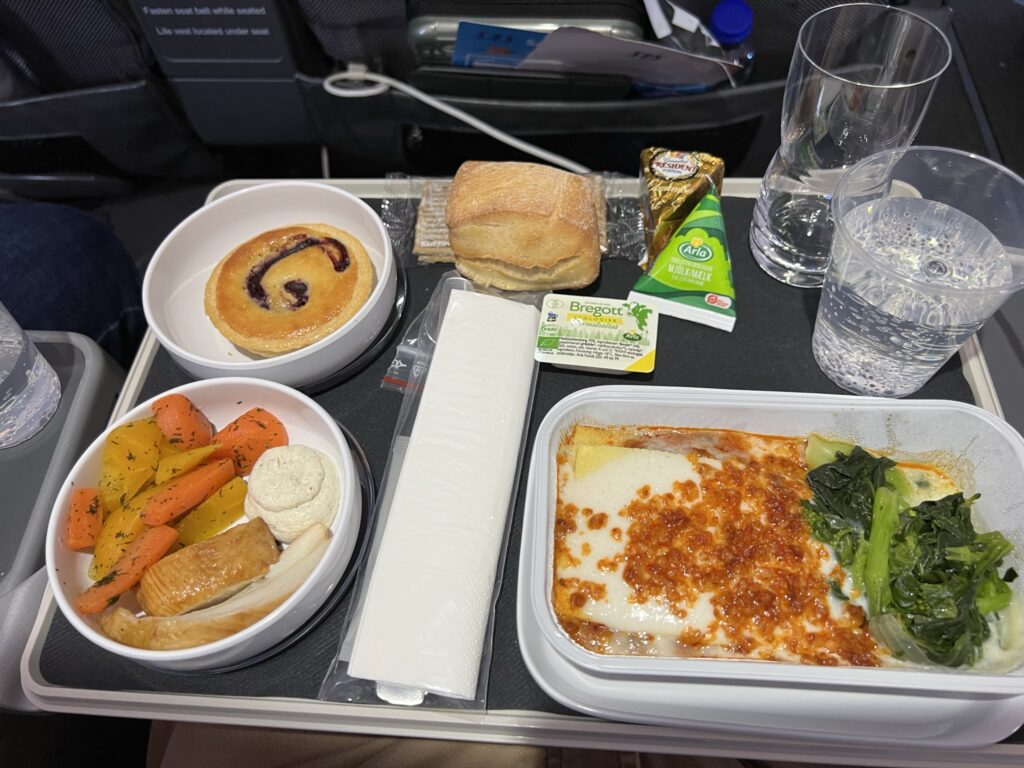 Inflight lasagna meal displayed on the aircraft tray table with some other snacks and a desert. 