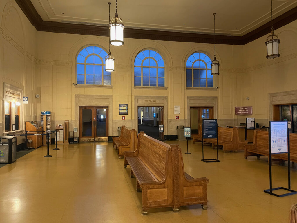 A large open train hall with old wooden benches for seating and various signs for information. 