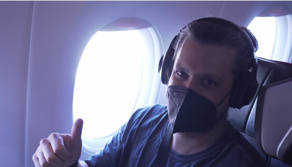 A masked male passenger is giving the thumbs up in premium economy while wearing headphones.