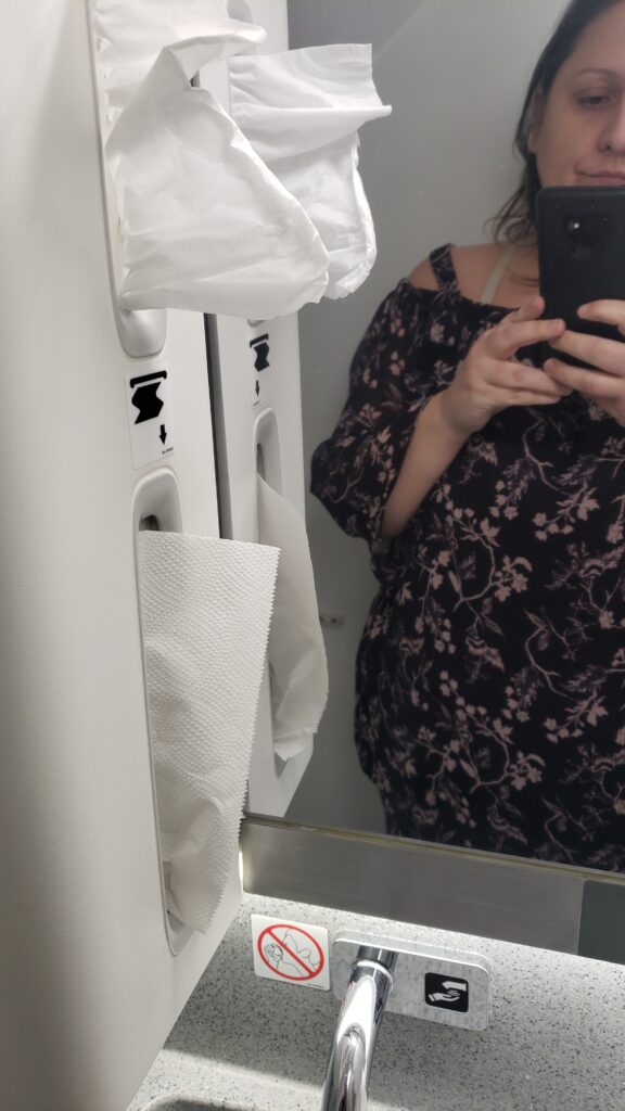 The author, a female passenger, is taking a selfie in the aircraft lavatory. The sink and paper towel/tissue compartment is in view. It appears to be a tight space.