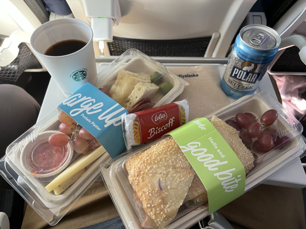 A packaged sandwhich, cheese platter, soda water and coffee sit on the aircraft tray table. 