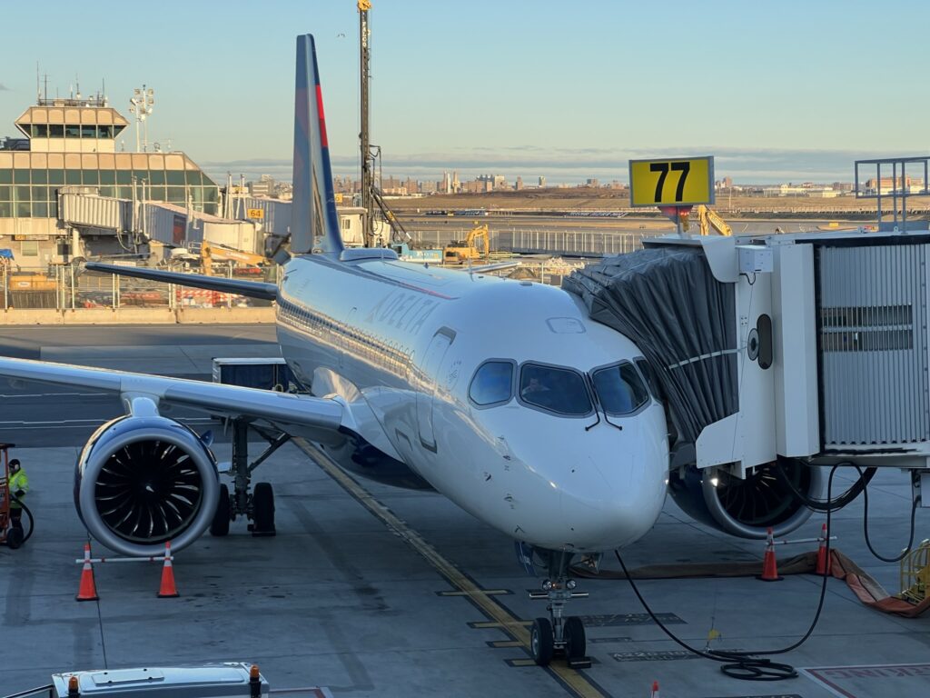 Delta A220 at the gate at LaGuardia on a sunny day. Ramp equipment and buildings can be seen.