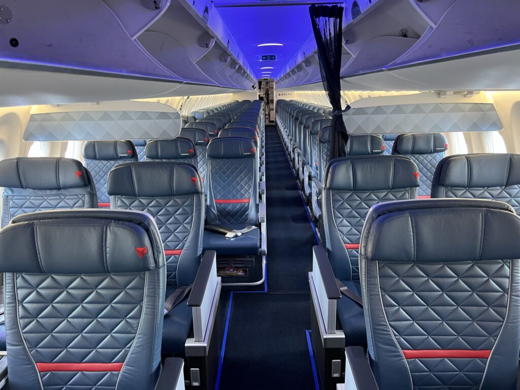 Delta Air Lines A220-100 cabin interior shows all navy blue seats with the signature red Delta accents. Blue LED lights shine from above the overhead bins. This image shows both first and main cabin seating. Large overhead bins are open, waiting for passengers.