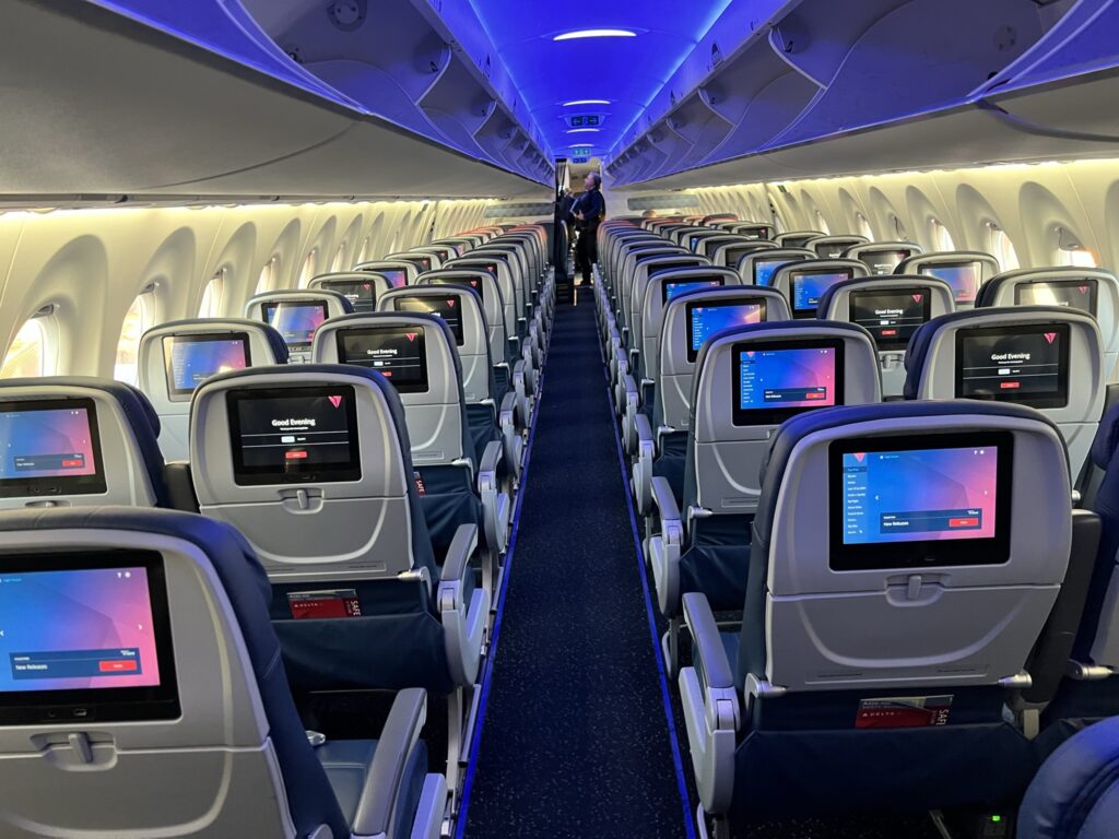 A photo taken aft of the cabin, looking up, shows rows of economy class seats with their seatback IFE screens turned on.