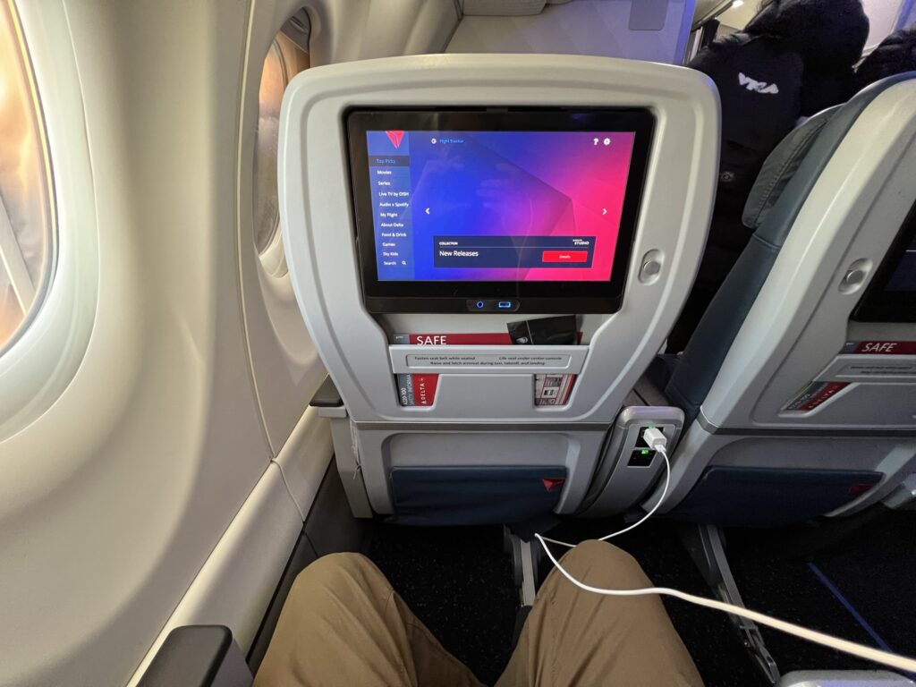 Delta Air Lines' A220-100 embedded IFE is showing the home screen. A man's knees are visible and a device cord is plugged into the inseat power.