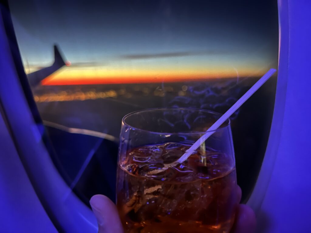 A drink is being held up in front of the aircraft window. Looking out of a city of lights at sunset.