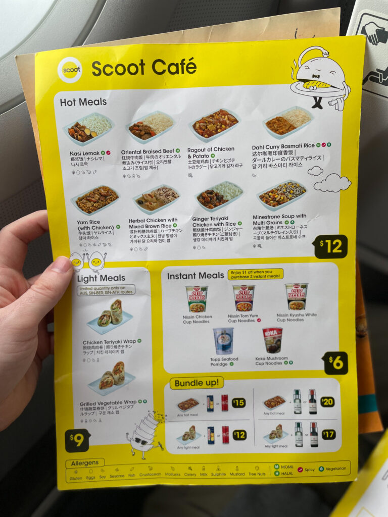 A passenger is holding up the yellow Scoot Cafe menu, which features various hot meals, instant meals and bundles with beverages.