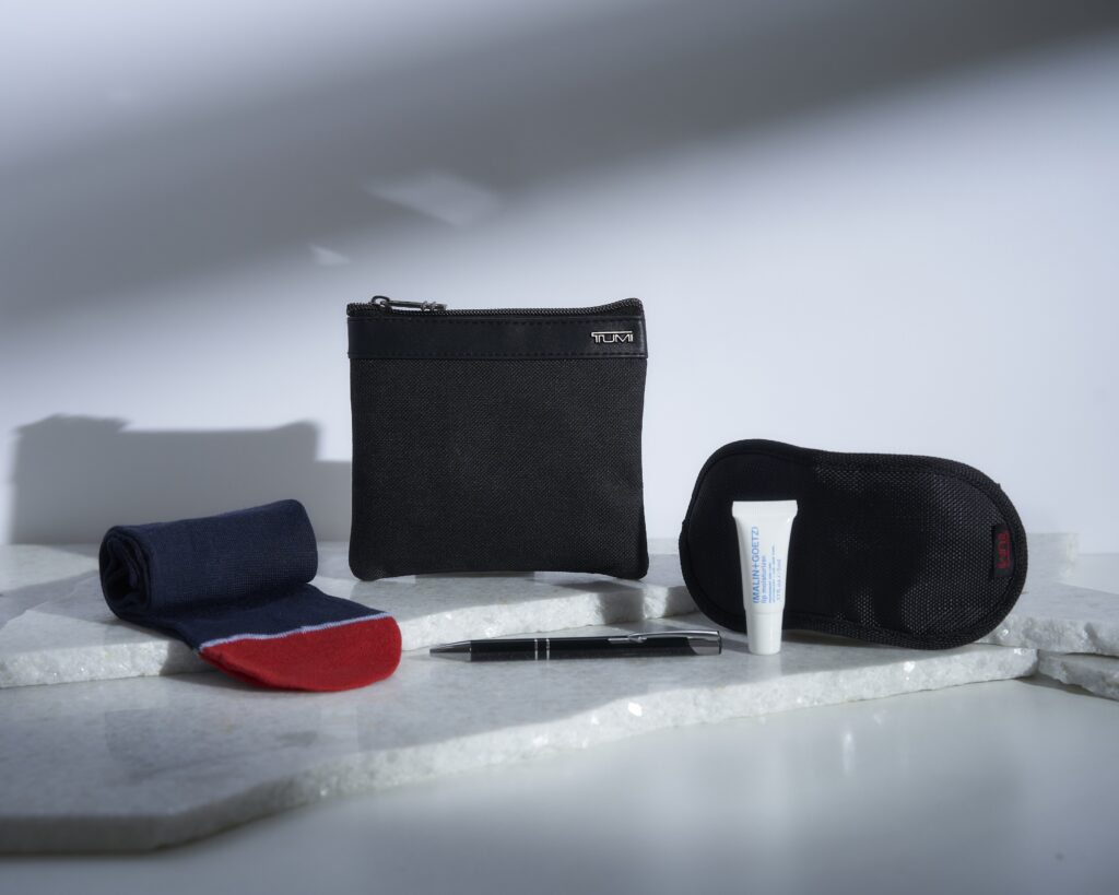 Blue and red socks, a pen, hand cream, black sleep mask and a black zip up pouch to hold it all is displayed as Air India TUMI Premium Economy Kit by Buzz.