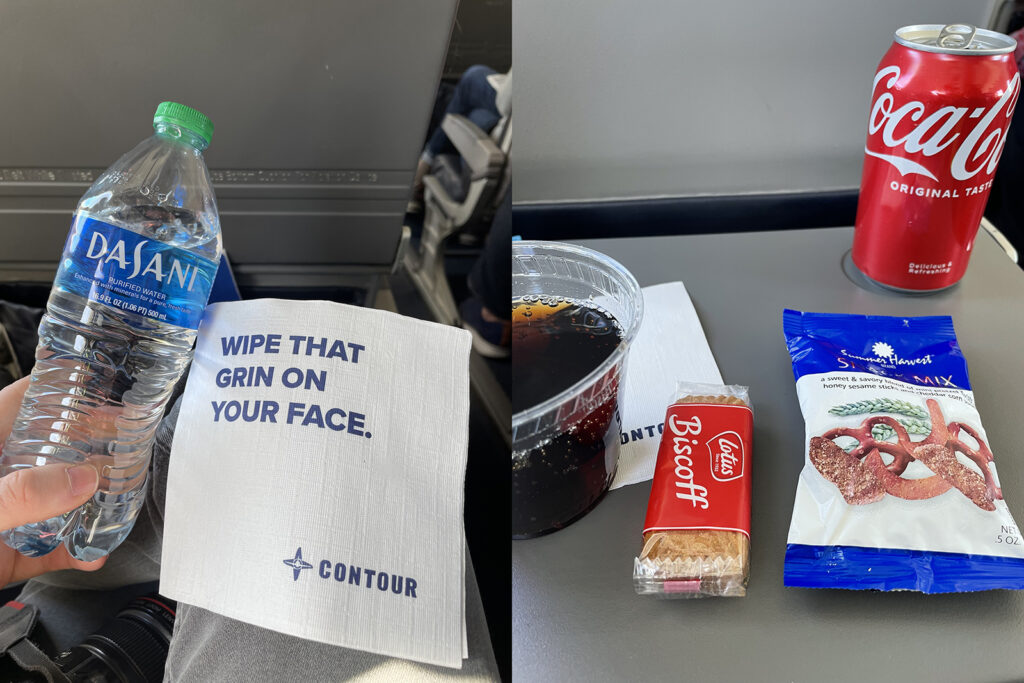 Snacks and drinks on the tray table. The author holds up a large bottle of Dasani water. A napkin reads: "Wipe that grin on your face."