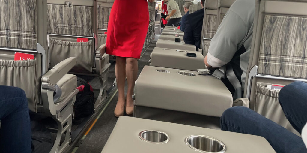 The JSX ERJ-145 has a side table with cup holders. A flight attendant, dressed in red, walks down the aisle.