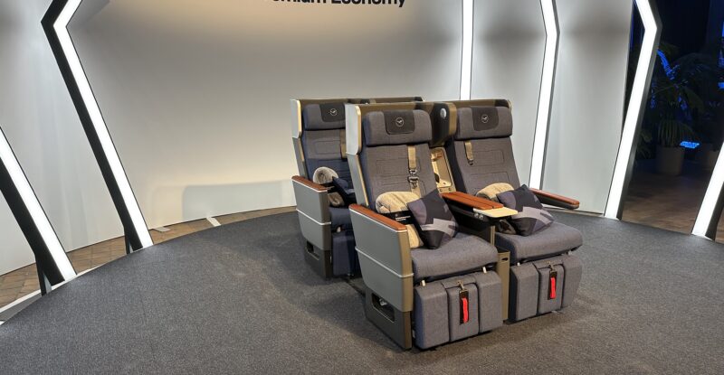 The Lufthansa Allegris premium economy class seat is on display. Thter are four seats displayed in Navy blue with wood finishings.